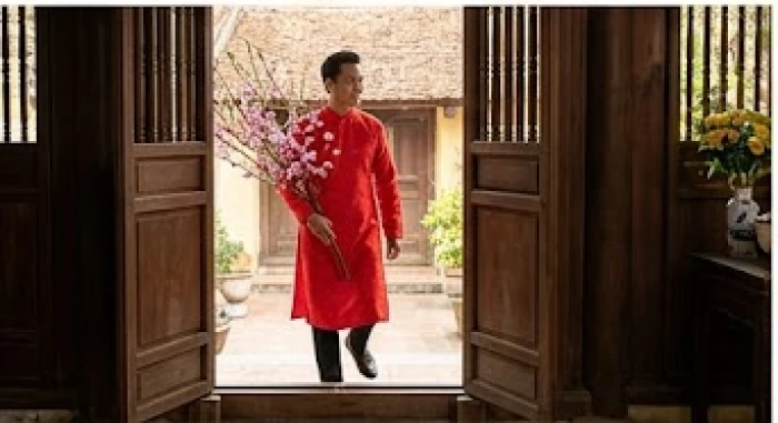 Người khách nam mặc áo dài đỏ cầm cành đào tươi thắm bước qua bậu cửa ngày đầu năm